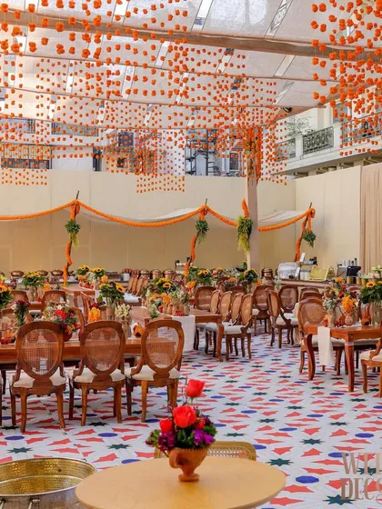 A wide shot of the dining area under a canopy of hanging marigold buds. The custom-printed geometric floor in red, white, and blue adds a contemporary touch to the traditional Indian decor elements.