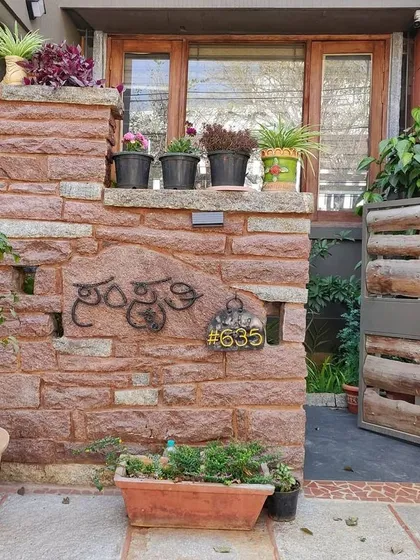 The entrance to a home named 'Samskruthi'. The rough-hewn stone compound wall and a gate made of unfinished wood logs create a rustic and earthy welcome, grounded in natural textures.