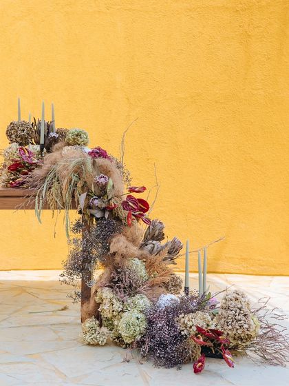 An editorial shot of a unique floral installation, featuring dried grasses and flowers against a bold yellow wall.