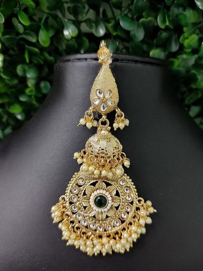 A large, circular maang tikka with a jhumka drop, featuring a green stone in the center.