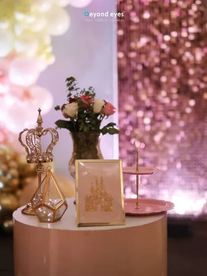 A close-up of the elegant decor details from a Disney Princess party, including a miniature golden crown and a framed art print, set against a sparkling backdrop.