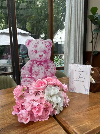 A charming table centerpiece for a Dior-themed baby shower, featuring a pink floral arrangement and a custom-printed teddy bear prop.