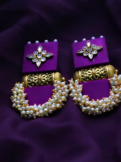 A detailed product shot of handmade purple and gold earrings against a matching purple fabric, emphasizing their color and intricate beadwork.