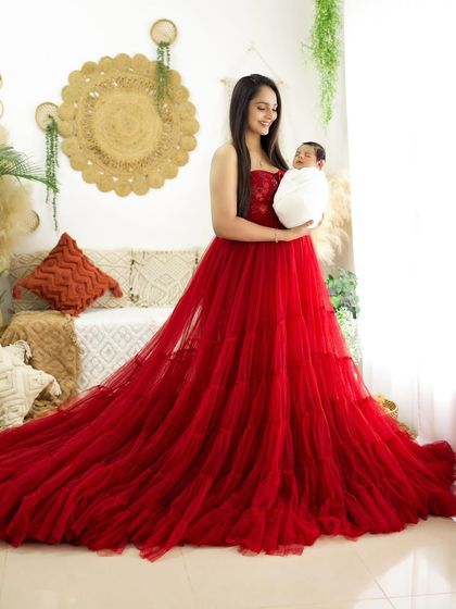 A wider shot of the mother and newborn, showcasing the full, flowing red gown against the bright and airy boho studio setup.