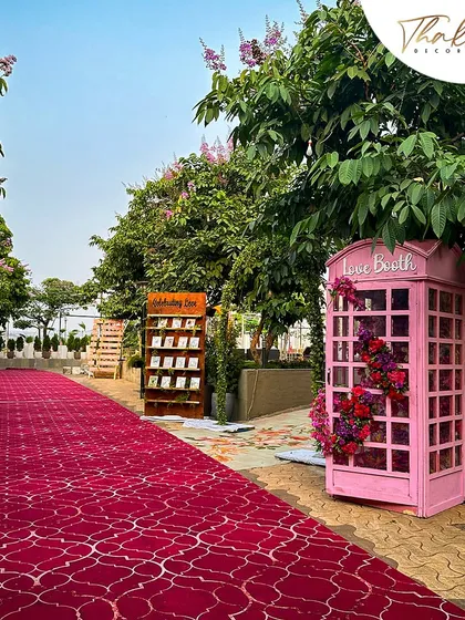 A beautiful outdoor wedding entrance featuring a red carpet, a 'Love Booth' photo op, and a wooden stand displaying the couple's photos, creating a personalized journey for guests.