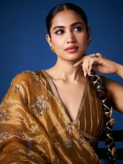 A close-up on the model's confident gaze, with the golden organza saree framing her face. The intricate embroidery and v-neck blouse are visible in detail.