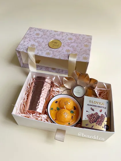 A unique hamper featuring a bowl of motichoor laddoo candles, a copper tumbler, and almond brittle, all presented in a beautiful handled box.