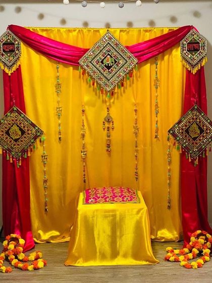 This compact Haldi setup is perfect for smaller indoor spaces. It combines a rich yellow backdrop with contrasting pink drapes and decorated with traditional embroidered hangings for a complete and festive look.