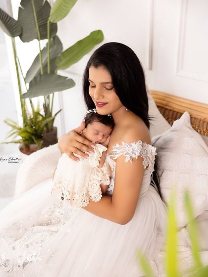 A mother cradling her newborn daughter, both dressed in beautiful white lace. The natural light from the window adds a soft, ethereal glow.