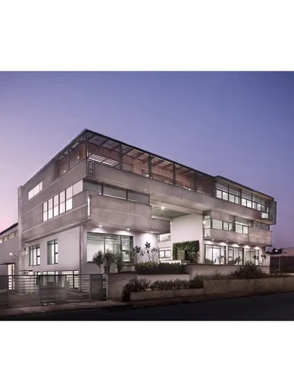 The corporate office seen at twilight, where the folding concrete planes of the facade are highlighted. The central entry court, connected to the street by landscaped mounds, creates an intriguing experience of raw concrete contrasting with tropical greenery.