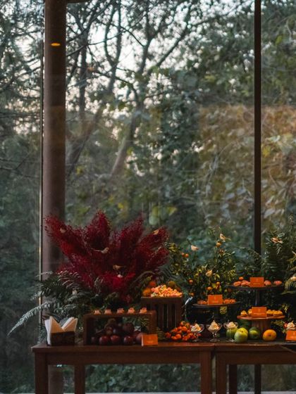 The vibrant red floral arrangements on this grazing table create a dramatic contrast with the lush green forest visible through the window, blending nature with culinary art.