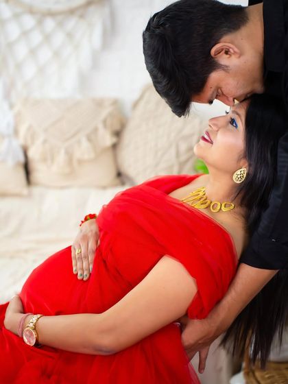 An intimate and loving pose. The partner leans in to kiss the mom-to-be's forehead as she reclines in a beautiful red gown, creating a romantic and tender portrait.