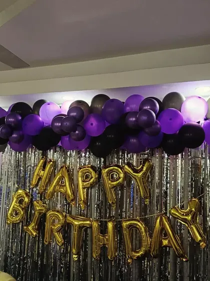 A purple and black balloon garland over a silver tinsel curtain with gold 'Happy Birthday' foil letters.