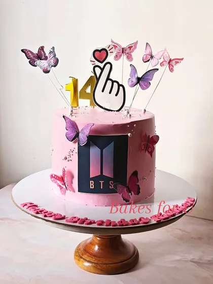 A BTS Army cake for a 14th birthday, decorated in the band's signature pink and black colors. The design includes the BTS logo, butterflies, and the iconic finger heart gesture.