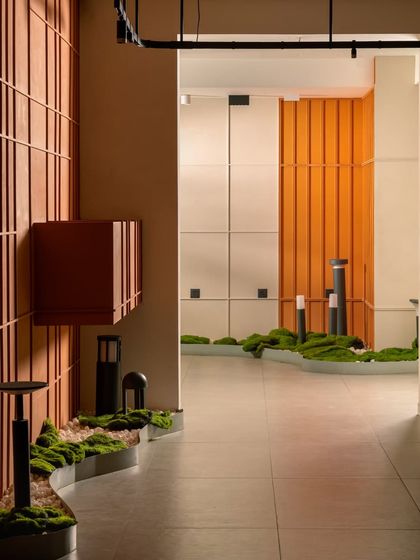 This angle showcases the layered design, with the terracotta grid wall in the foreground and the bright orange paneling in the distance. The outdoor lighting fixtures are displayed naturally along the moss bed.