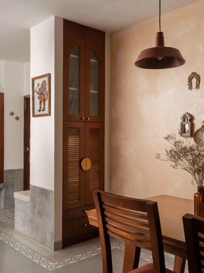 The dining area is defined by a custom teak crockery unit and a warm, textured wall finish. The design carves out an intimate dining space while maintaining a connection to the living area.