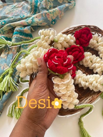 A close-up of my desi-inspired crochet gajra. I use off-white yarn to mimic mogra flowers and add pops of red with tiny roses, all held together with green yarn tassels.
