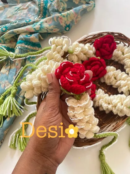 A close-up of my desi-inspired crochet gajra. I use off-white yarn to mimic mogra flowers and add pops of red with tiny roses, all held together with green yarn tassels.