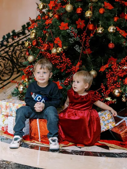 Two siblings sitting patiently by the Christmas tree, surrounded by presents. A classic and beautiful holiday portrait.