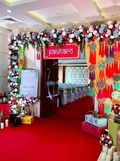 A festive and colorful entrance featuring a 'Padharo' sign, dreamcatcher decorations, and a floral arch, all leading down a vibrant red carpet.