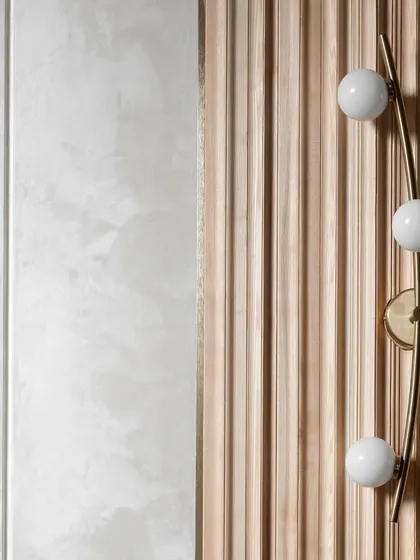 A study in texture and light. This composition features a modern brass wall sconce with globe lights set against a fluted wooden panel, which contrasts with the smooth, lime-plastered wall beside it.