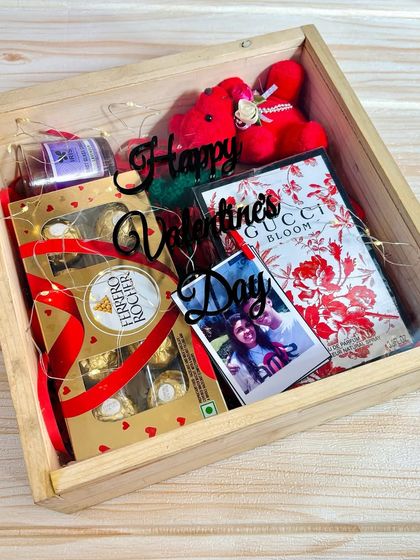 A top-down view of the romantic wooden box hamper, showing the "Happy Valentine's Day" acrylic cutout and the carefully arranged gifts, including perfume and a personal photo.