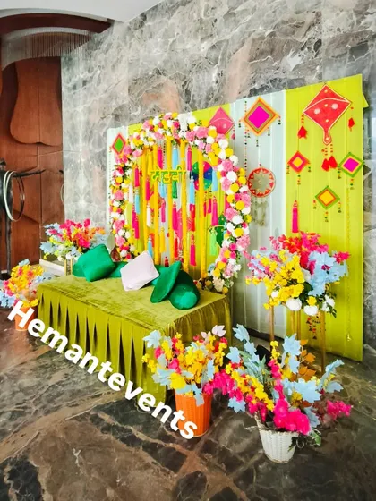 An indoor Mehendi or Haldi corner setup against a marble wall. The bright green and yellow backdrop is decorated with kites and floral arrangements for a festive look.