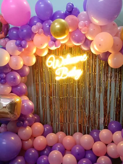 This 'Welcome Baby' decoration uses a gold shimmer curtain as a backdrop to create a festive look. The scene is framed by a balloon arch in purple, pink, and gold, with a central neon sign.