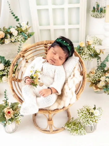 A beautiful overhead shot of a baby sleeping in a round wicker chair, surrounded by delicate flowers. The green headband adds a lovely pop of color.