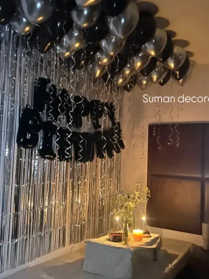 A romantic birthday surprise in a bedroom using black and silver balloons. The setup includes a silver tinsel curtain and a small table for a cake, creating an intimate celebration space.