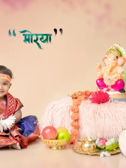 A beautiful Ganpati festival photoshoot featuring a baby girl in a traditional blue and red outfit. The setup is complete with a Ganpati idol, fruits, a dholak drum, and festive decorations, capturing the devotional spirit of the occasion.
