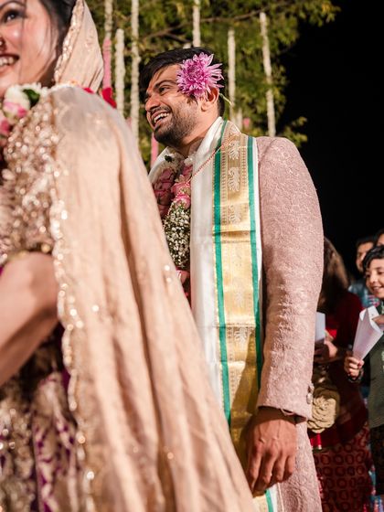 The groom beaming during the wedding ceremony, a flower tucked in his hair, his pink sherwani perfectly complementing the bride's look.