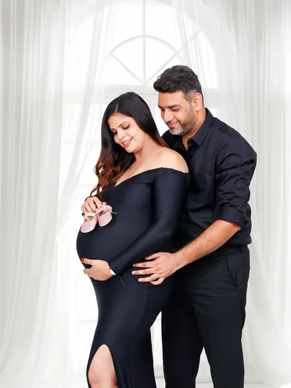 An elegant and classic studio portrait of an expecting couple. Dressed in matching black, they share a tender moment as they both hold the baby bump, with tiny pink baby shoes hinting at the little one's arrival.