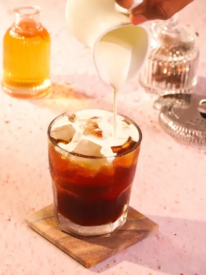An action shot of cream being poured into a glass of iced coffee. The pink-toned lighting adds a unique and warm glow, highlighting the swirl of the cream as it mixes with the dark coffee.