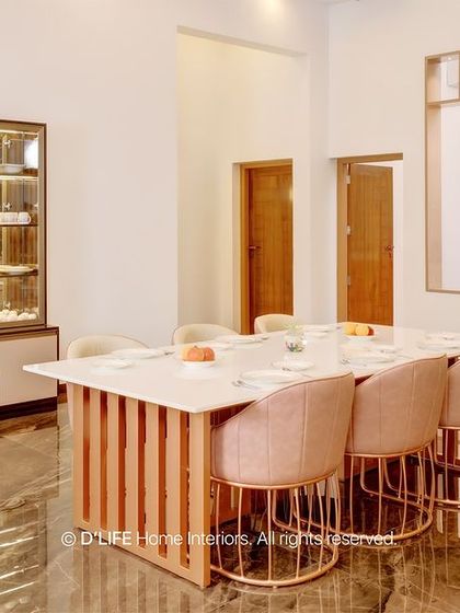 A luxurious dining room featuring a marble-topped table with a fluted rose-gold base, paired with pink upholstered chairs. A large glass-front crockery unit provides elegant storage.