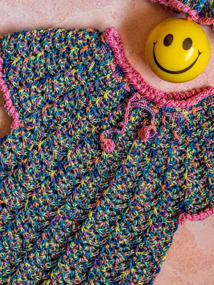 This handmade crochet dress is a burst of color and texture. The vibrant, multi-colored yarn is complemented by a simple pink trim, making it a truly unique and eye-catching piece.