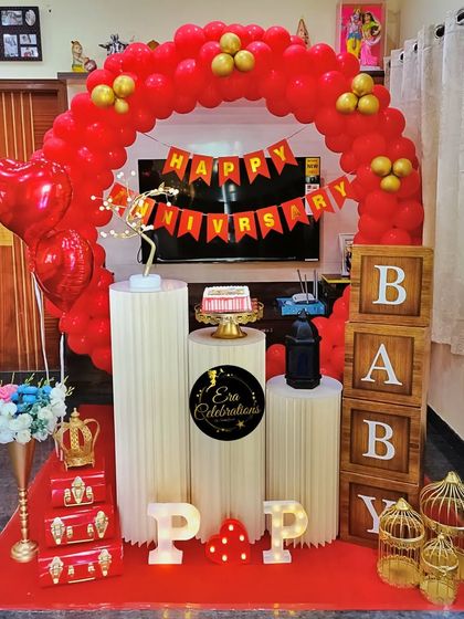 A vibrant red and gold anniversary decoration for a home celebration. The setup includes a red balloon arch, heart-shaped balloons, and decorative props.