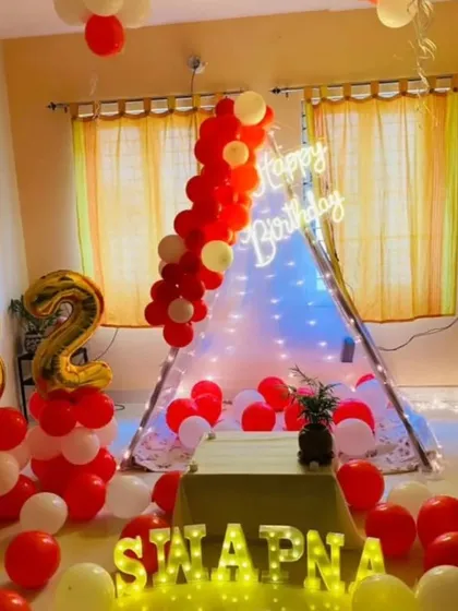 A cozy and cute birthday setup featuring a canopy tent with fairy lights, red and white balloons, and the birthday person's name in marquee letters.