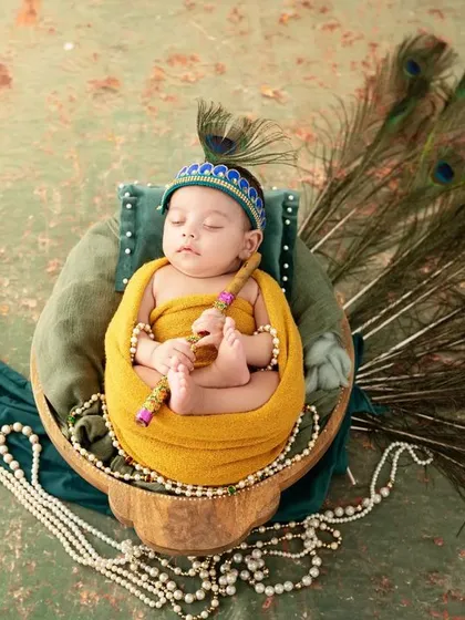 A creative and cultural newborn portrait of a baby dressed as Little Krishna. The baby rests in a basket designed to look like a peacock, complete with a full feather tail, holding a tiny flute.