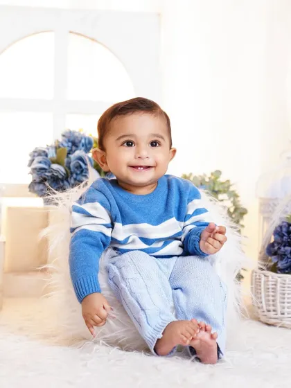 A happy baby boy dressed in a cute blue sweater, sitting in a bright, airy studio setup with blue floral decorations.