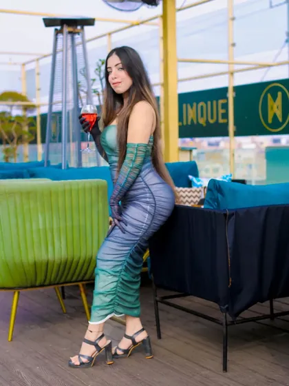 A full-length shot that captures the glamour of the outfit and the location. The pose is confident and powerful, creating a high-fashion image in a lounge setting.