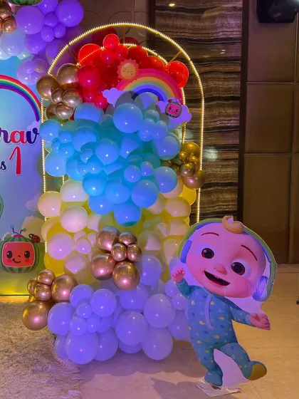 A vibrant Cocomelon theme for a first birthday party. This setup includes a custom arched backdrop, a rainbow-colored balloon arrangement, and a life-sized cutout of the character JJ.