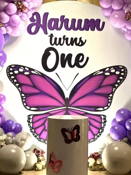 A close-up of the butterfly themed backdrop, focusing on the "Harum turns One" text and the large, vibrant purple butterfly at the center.