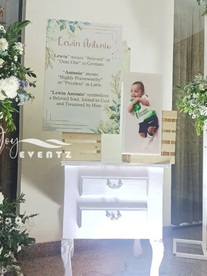 A personalized entrance display for a baptism, featuring a photo of the child and a sign explaining the meaning of their name. The decor is accented with a vintage-style table and fresh flowers.
