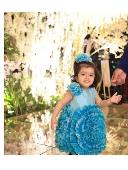 A vision in blue. This custom flower ball dress features a skirt made of countless handcrafted organza ruffles, creating a stunning, voluminous look. It’s paired with a matching floral headband for a picture-perfect first birthday outfit.