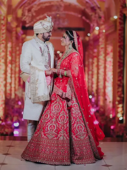 A stunning portrait of the couple in a grand, beautifully lit corridor. The bride's red lehenga and the groom's ivory sherwani create a classic and royal look.