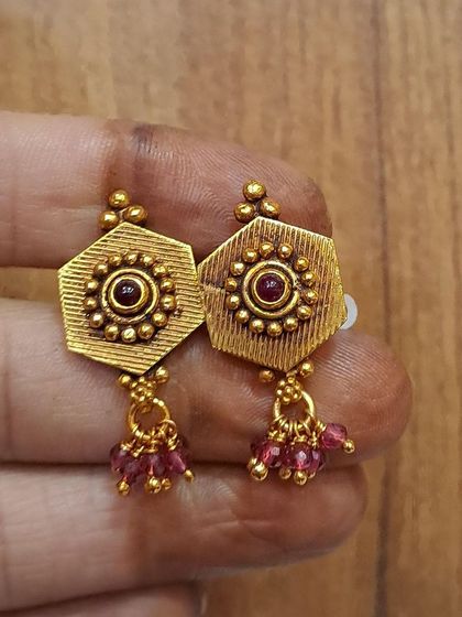These hexagonal antique gold-plated earrings are adorned with a central stone and ruby-colored bead drops. They are a beautiful blend of geometric and traditional design.