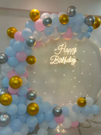 A pastel blue, pink, white, and gold balloon arch against a sheer curtain with fairy lights.