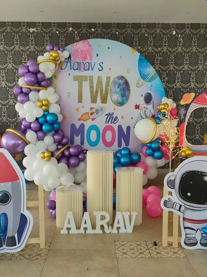 A "Two the Moon" space theme for a second birthday. This setup includes a circular galaxy backdrop, astronaut and rocket cutouts, and a purple and blue balloon garland.