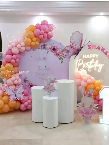 A bunny and butterfly theme for a first birthday. This decoration features a custom backdrop, a pink and orange balloon arch, and a marquee number '1'.